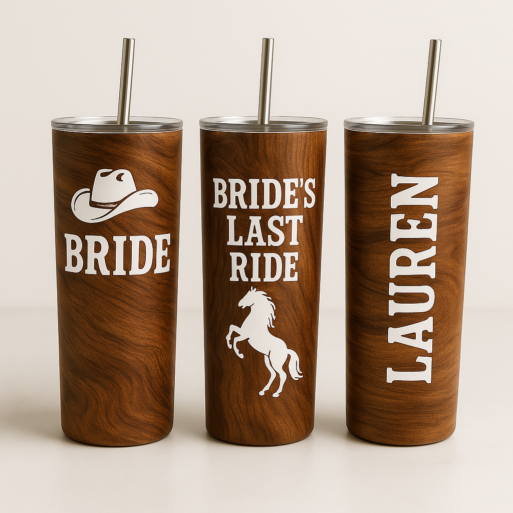 A set of three tall stainless steel tumblers with a rich wood-grain finish is displayed on a light surface. The first tumbler features a white cowgirl hat graphic with the word “BRIDE.” The center tumbler reads “BRIDE’S LAST RIDE” with a white rearing horse graphic. The third tumbler is personalized with the name “LAUREN” in bold white vertical lettering. Each tumbler has a clear lid and a stainless steel straw, creating a cohesive western bachelorette theme.